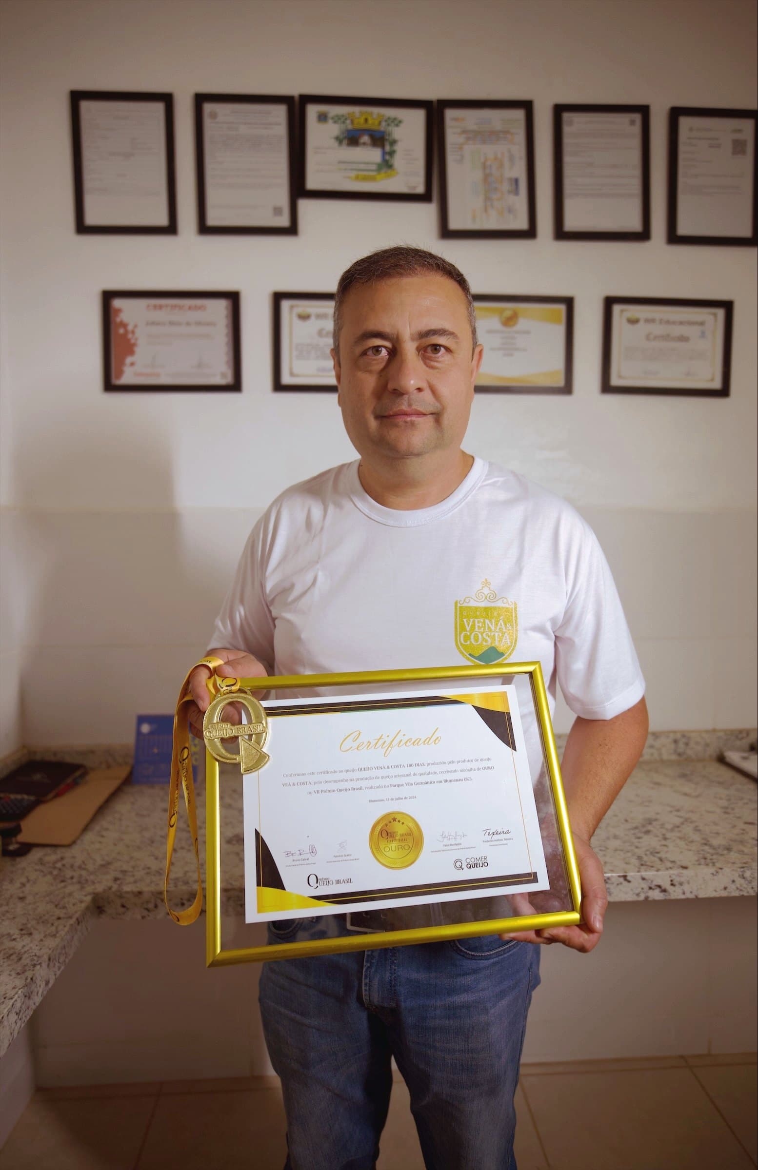 Juliano Oliveira segurando um certificado emoldurado na queijaria, com outros certificados na parede